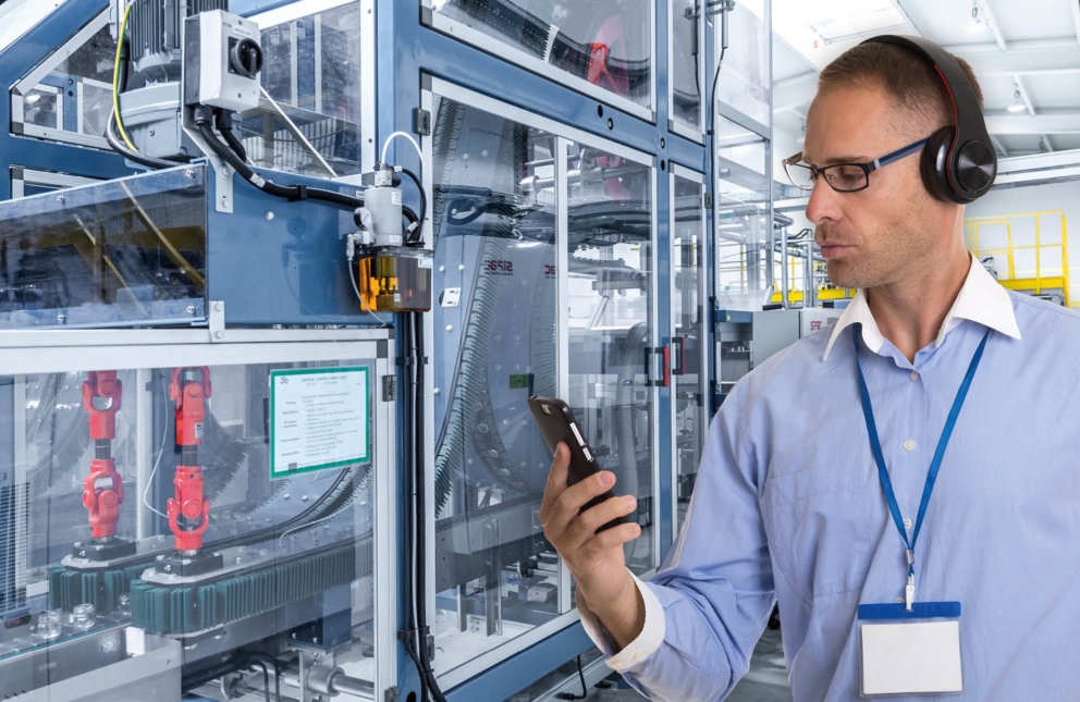 Eine Person in der Fabrikhalle hält ein Smartphone und trägt Kopfhöhrer