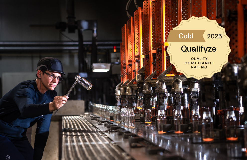 A factory worker wearing protective gear inspects a glass bottle on a production line. Overlay shows a "Gold 2025 Qualifyze Quality Compliance Rating" badge.